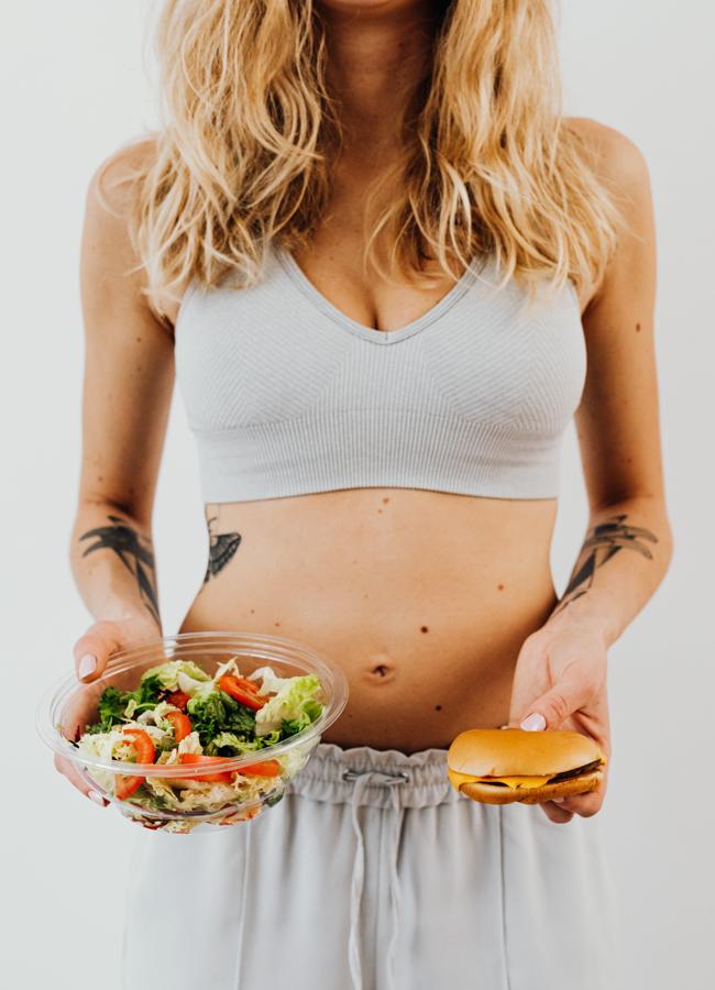 Mujer joven decidiendo entre comer una ensalada o una hamburguesa. / Foto de Karolina Grabowska en Pexels.