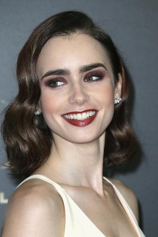 Lily Collins en los Hollywood Film Awards con media melena peinada con ondas amplias retro y un maquillaje muy llamativo con labios rojos y ojos ahumados en tonos berenjena.